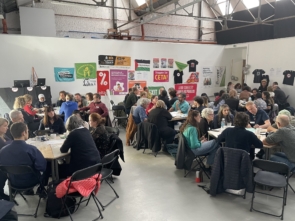 Many people sitting round tables in a warehouse style room. The back wall is covered in banners