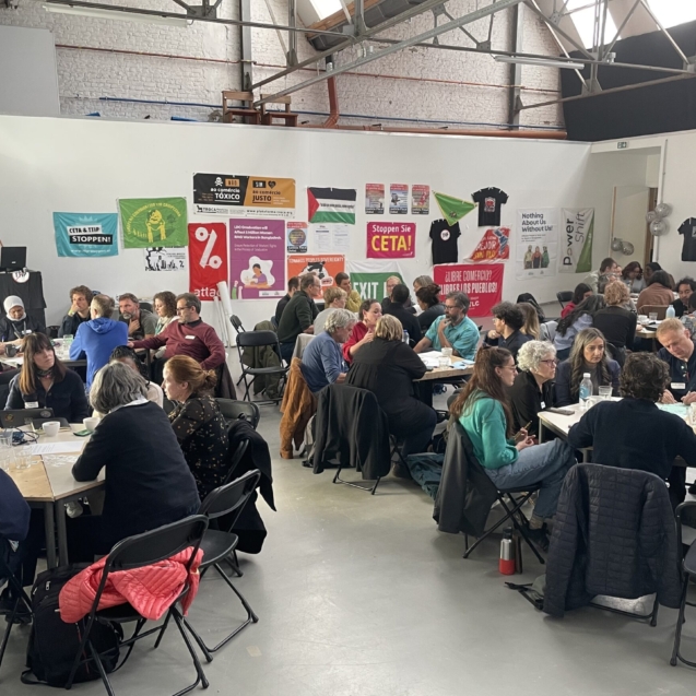 Many people sitting round tables in a warehouse style room. The back wall is covered in banners