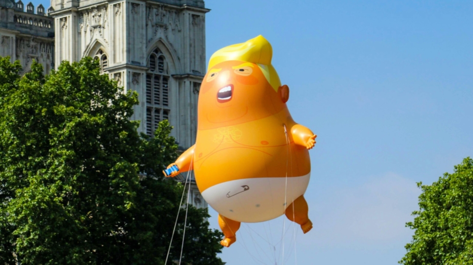 Giant inflatable shaped as a caricature of Donald Trump as a baby floats in front of some trees and a building